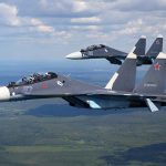 Two fighter jets flying in formation over green landscape.