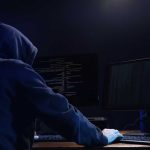Hooded figure working on computers in dark room