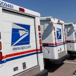Row of USPS mail trucks parked in lot.