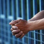 Hands holding blue prison bars tightly outside.