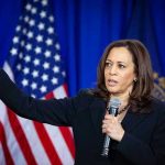 Woman speaking and gesturing with American flag background.