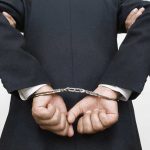 Person handcuffed in suit, escorted by two others.