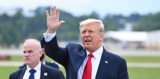 Man in suit waving while another man stands nearby.