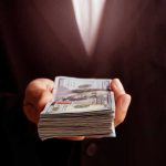 Your Taxpayer Cash Still Fueling Powerful Antisemitism Movement Hand holding a stack of hundred-dollar bills.