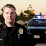 Public Stunned: Dangerous Arrest Leads to Officer’s Firing A police officer in uniform stands in front of a police car with lights on