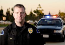 Public Stunned: Dangerous Arrest Leads to Officer’s Firing A police officer in uniform stands in front of a police car with lights on