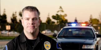 A police officer in uniform stands in front of a police car with lights on