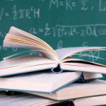 Math Proficiency Nosedive: Who’s to Blame? A stack of books with one open on top, in front of a chalkboard filled with mathematical equations