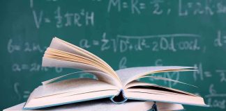 A stack of books with one open on top, in front of a chalkboard filled with mathematical equations