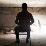 Person tied to chair in dimly lit room.