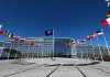 NATO Crumbles—Trump Forces Historic Deal Flags outside NATO headquarters building under clear blue sky.
