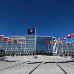 NATO Crumbles—Trump Forces Historic Deal Flags outside NATO headquarters building under clear blue sky.