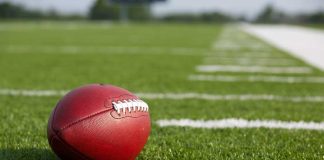 A red American football resting on a green artificial turf field