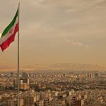 Iran’s Regime Surviving Massive Uprising Iranian flag waving over a city skyline with mountains in the background