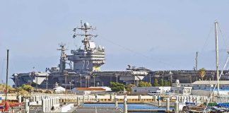 Trump’s Bold Move: Obama-Biden BLAMED! A large naval aircraft carrier docked in a harbor with smaller boats in the foreground