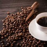 A cup of hot coffee next to spilled coffee beans and a wooden scoop