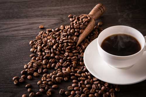 A cup of hot coffee next to spilled coffee beans and a wooden scoop