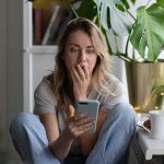 Woman sitting by plants looking at smartphone with surprised expression