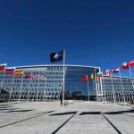 NATO HQ Infiltrated! — TRUSTED Intern Arrested NATO headquarters with member flags in front under a clear blue sky