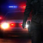 Police officer stands before patrol car with flashing lights at night