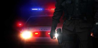 Police officer stands before patrol car with flashing lights at night