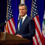 Man in suit speaking at a podium with American and California flags
