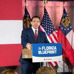Man in suit speaking at a podium with American and state flags behind