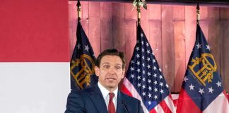 Man in suit speaking at a podium with American and state flags behind